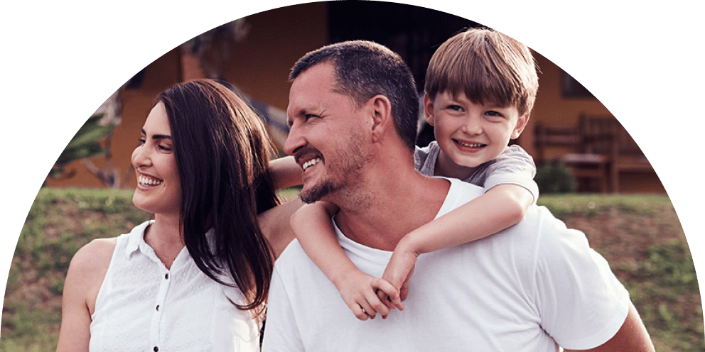 A happy family, a woman, man, and child, smiling outdoors. The boy is on the man's back.