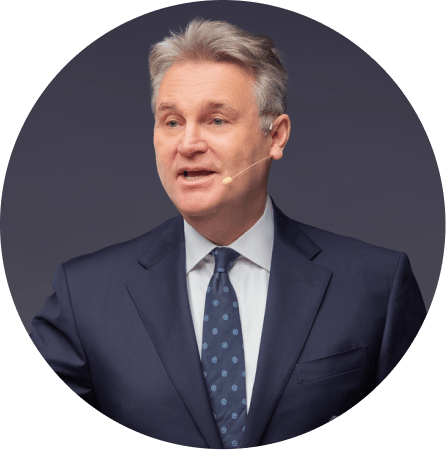 Man in suit and tie with grey hair speaking into an ear-worn microphone.