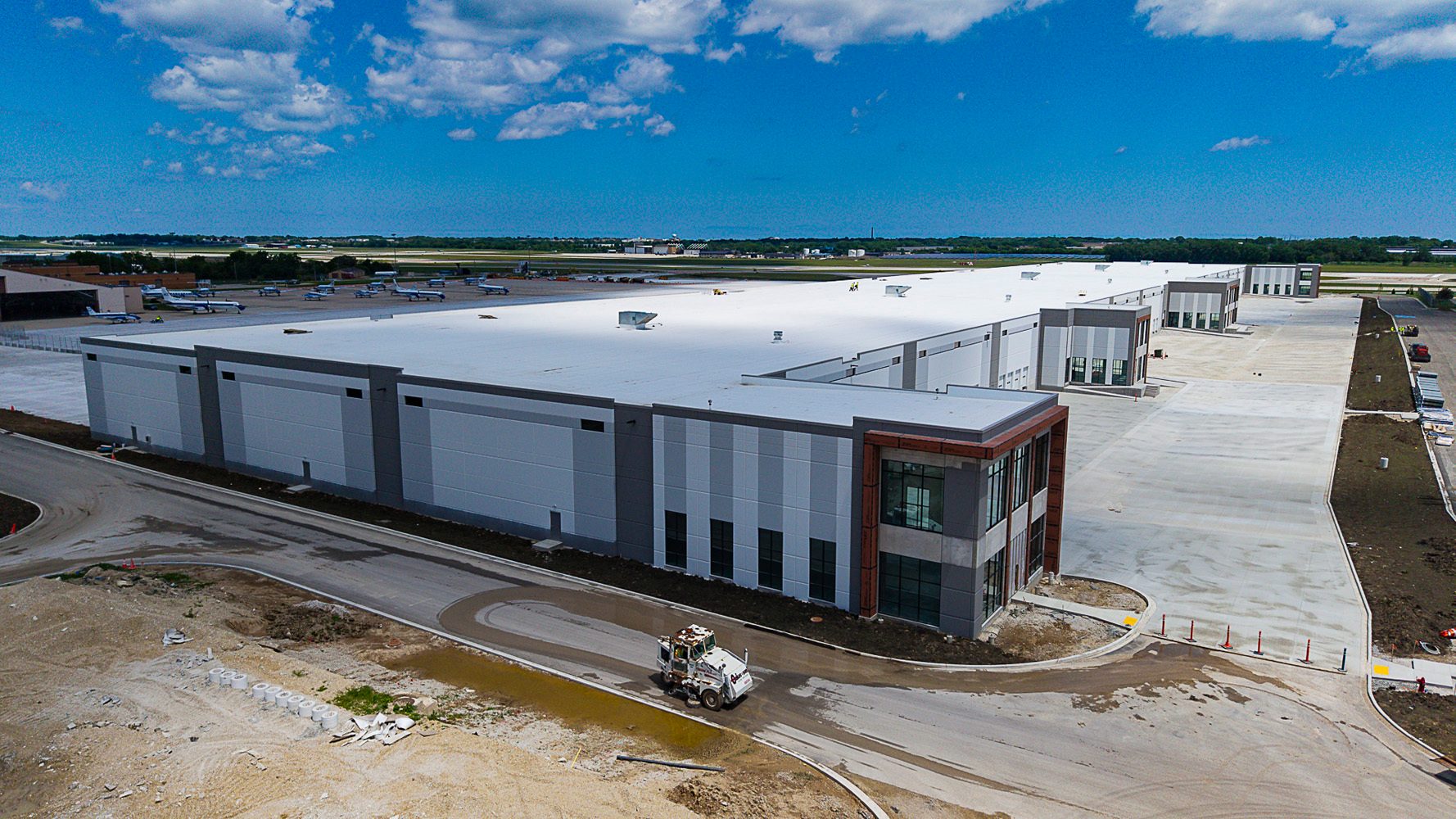 Large warehouse by airport tarmac with parked planes under blue sky, construction vehicle nearby.