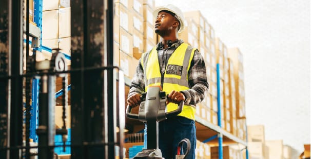 A Black man in a hard hat and safety vest operates a pallet jack in a warehouse with boxes.