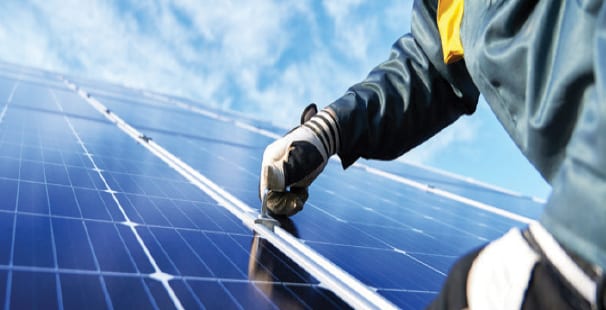 A gloved hand of a worker installing or maintaining a solar panel under a clear blue sky.