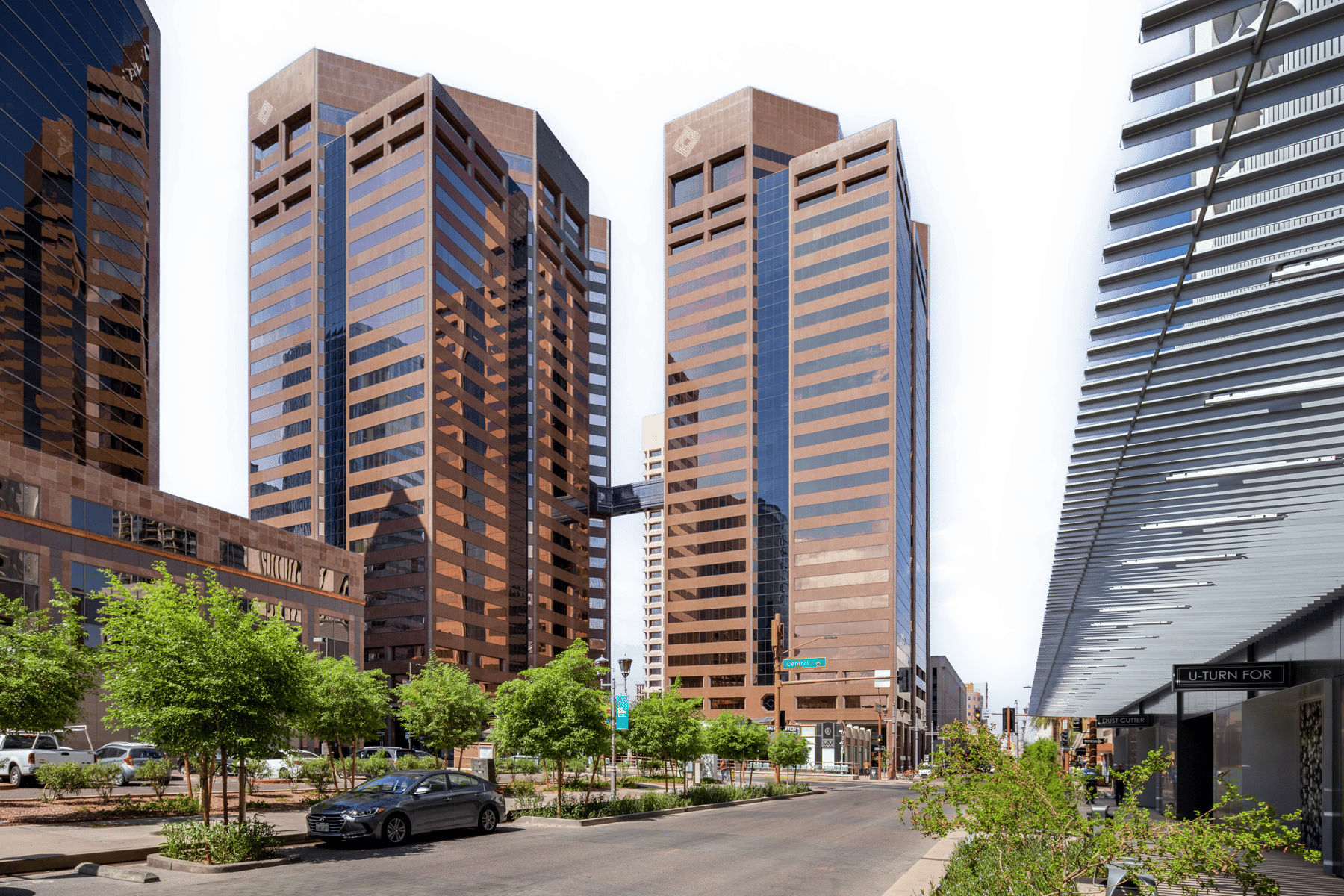 Tall brown office buildings and modern structures line an urban street with trees and a car.
