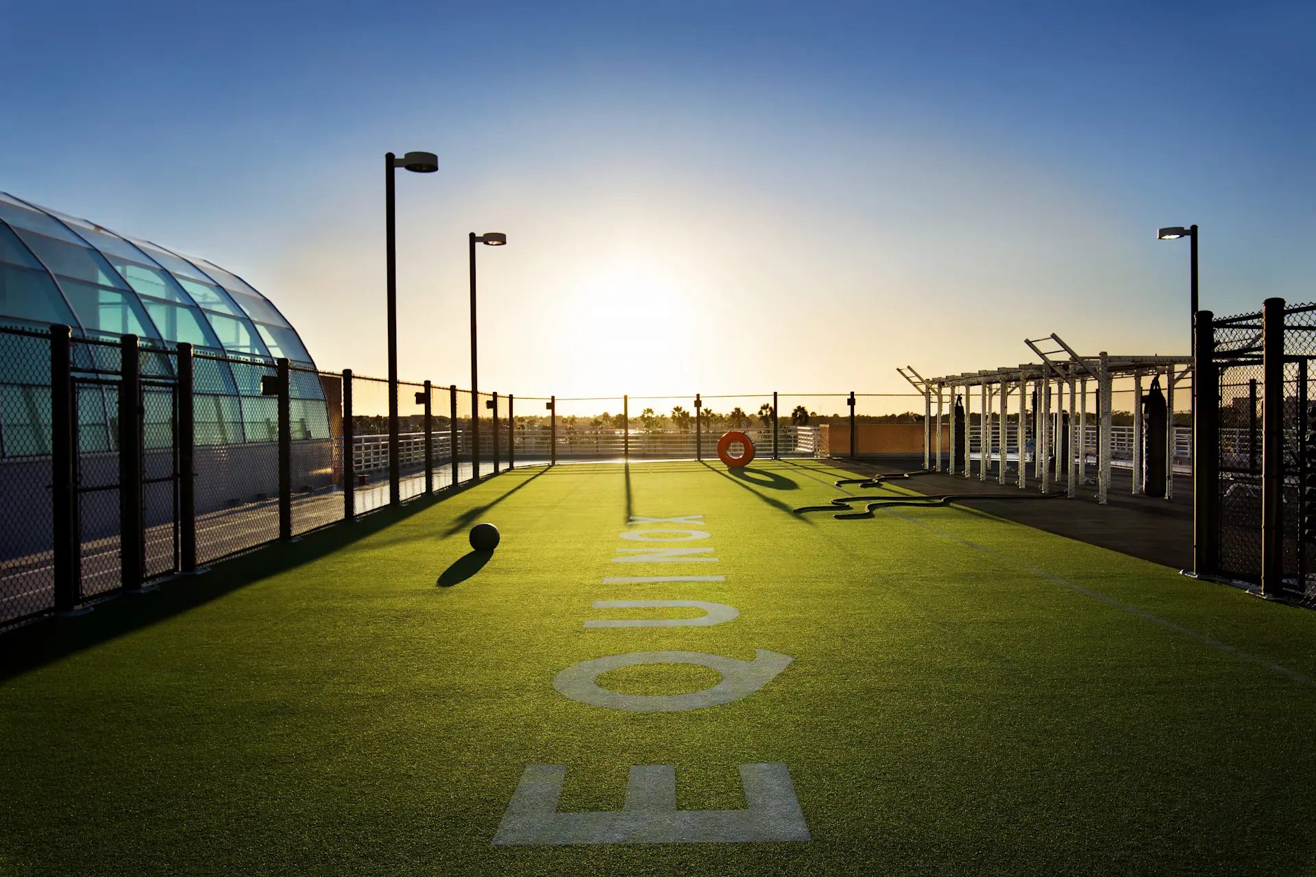 Rooftop fitness center with green turf, 'EQUINOX' branding, equipment, and a glass structure under a sunset sky.