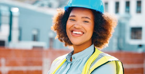 A smiling African American woman in a blue hard hat and yellow safety vest at a construction site.