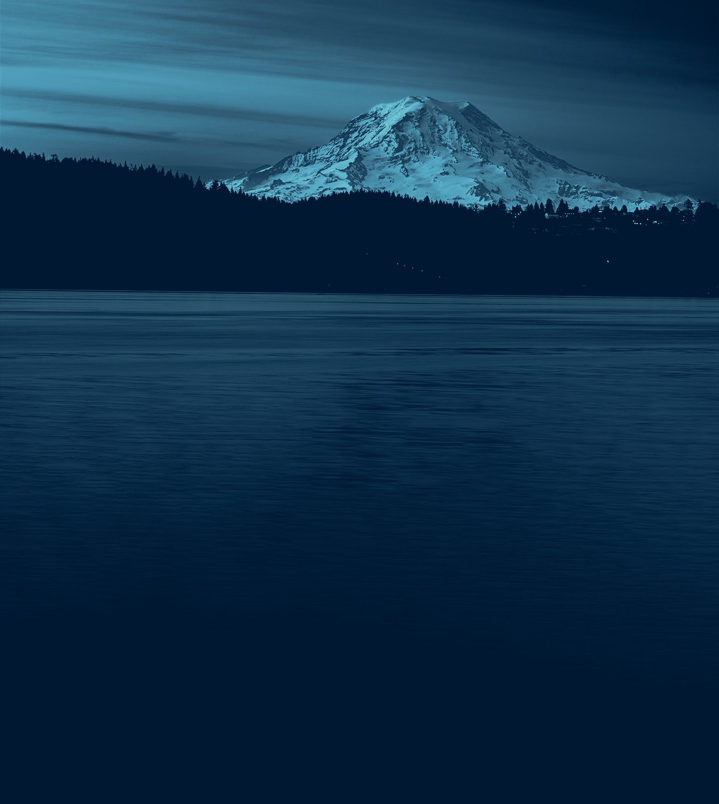 Snow-capped mountain over dark forest and deep blue lake under a twilight sky.