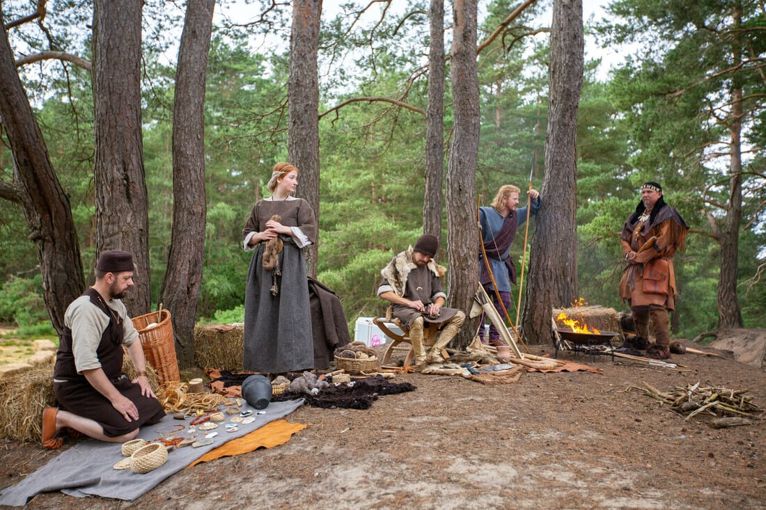 People in diverse historical attire demonstrating crafts in a forest camp.