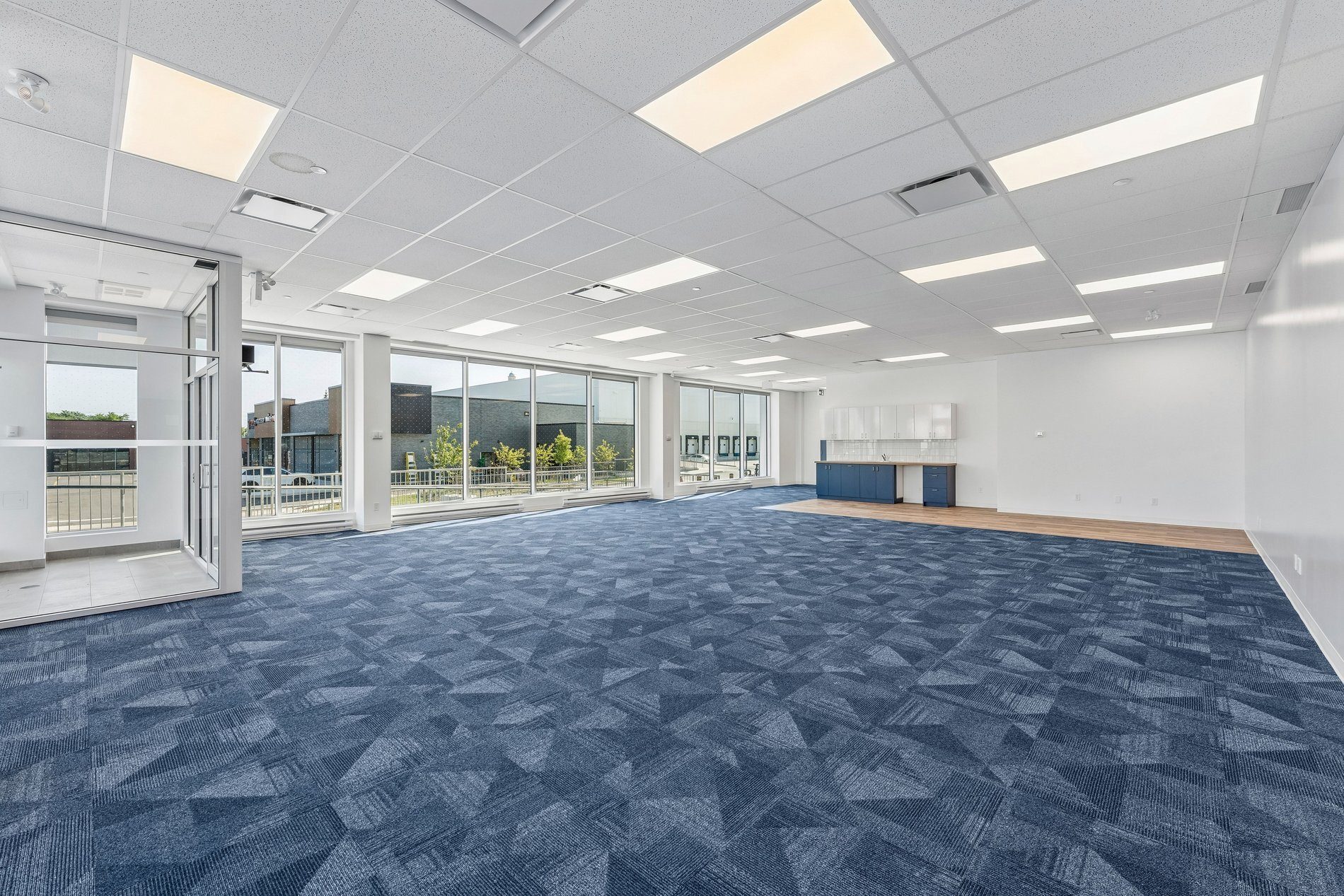 Spacious empty office with blue patterned carpet, large windows, white walls, and a small kitchenette.