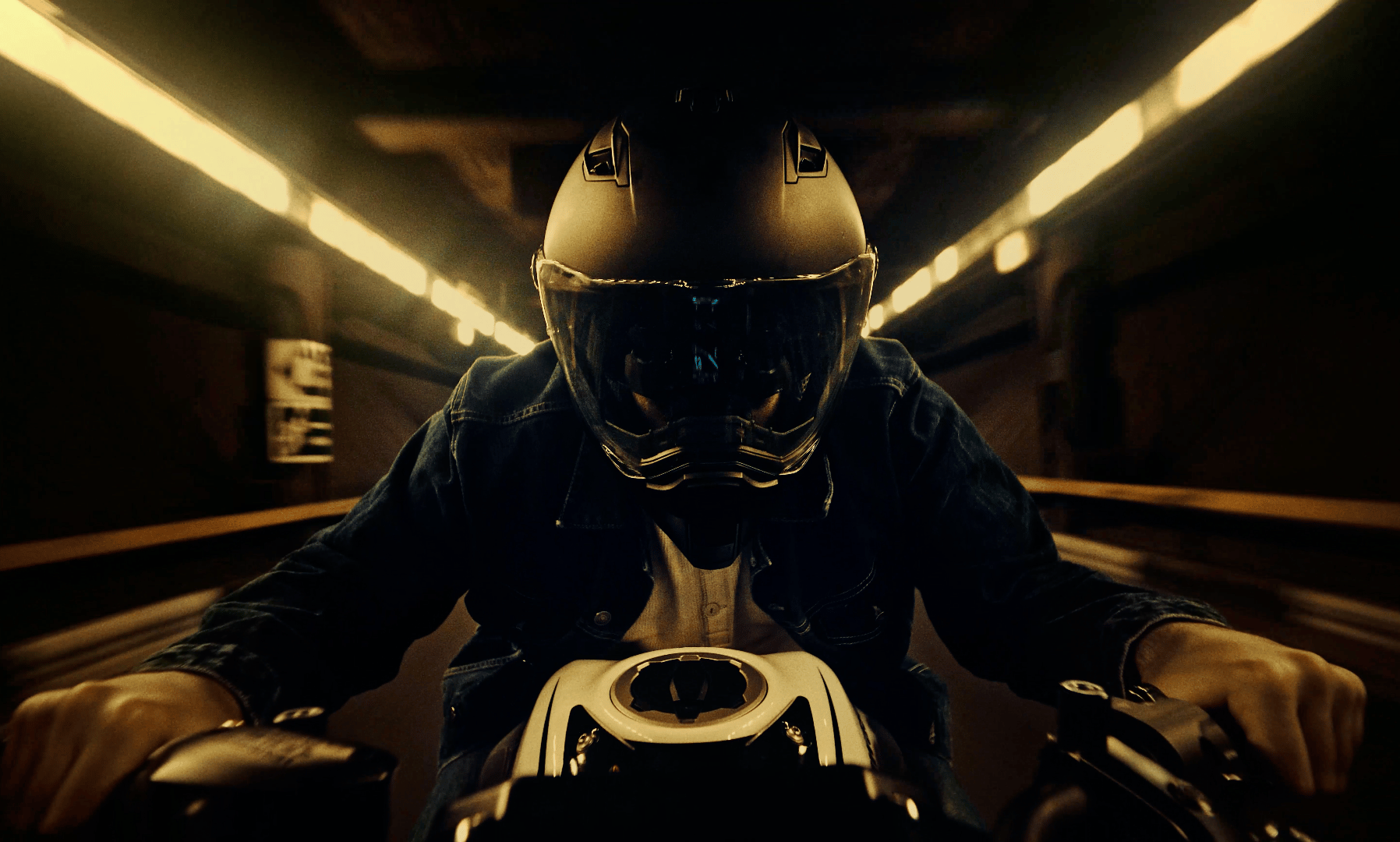 Motorcyclist in helmet in a dark tunnel, illuminated by bright strip lights.