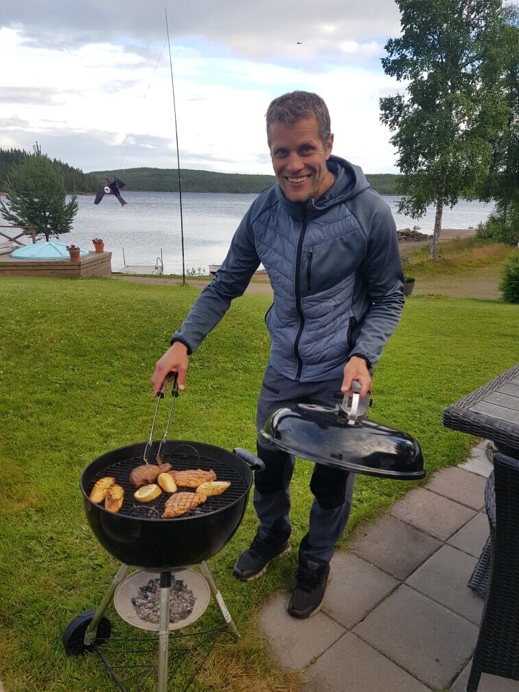A smiling man barbecuing food on a grill with tongs, with a scenic lake and trees in the background.