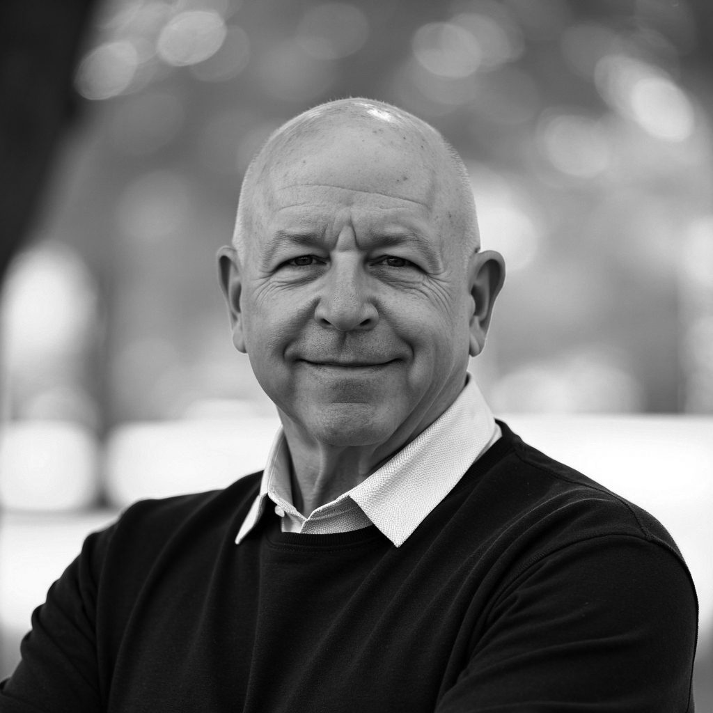 Black and white portrait of a smiling, bald man in a sweater and white collared shirt.