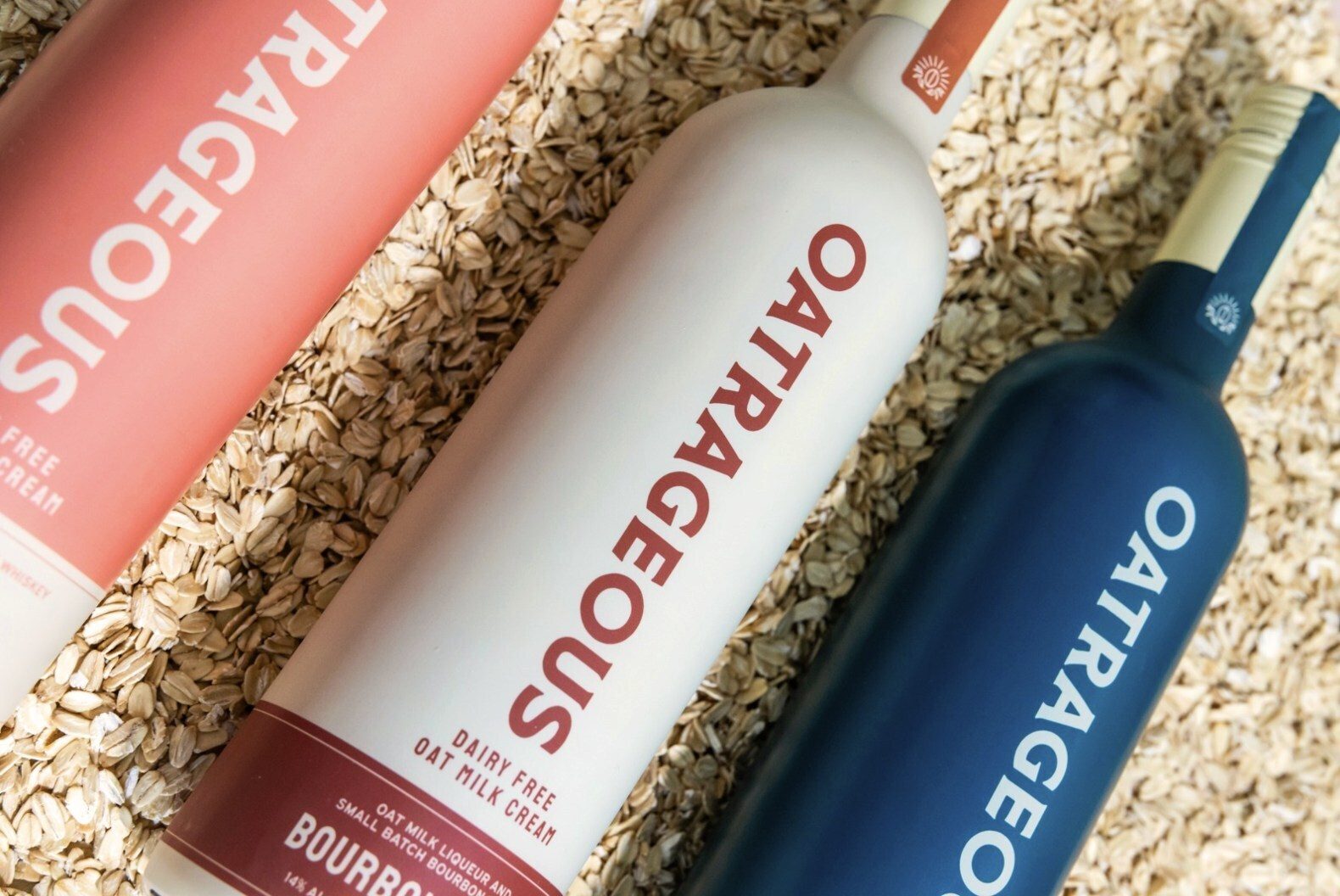 Three OATRAGEOUS oat milk liqueur bottles (pink, white, blue) resting on rolled oats.