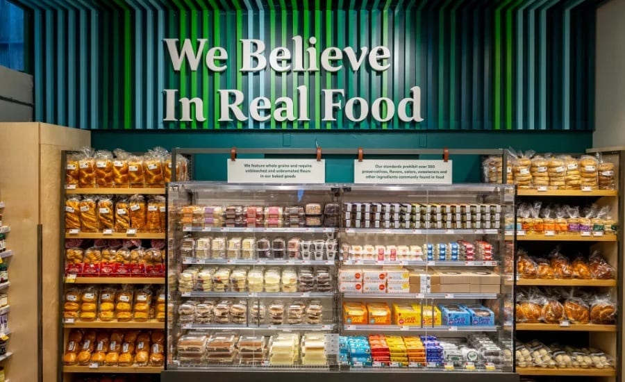 A grocery store aisle featuring baked goods and pre-packaged foods under a 'We Believe In Real Food' sign.