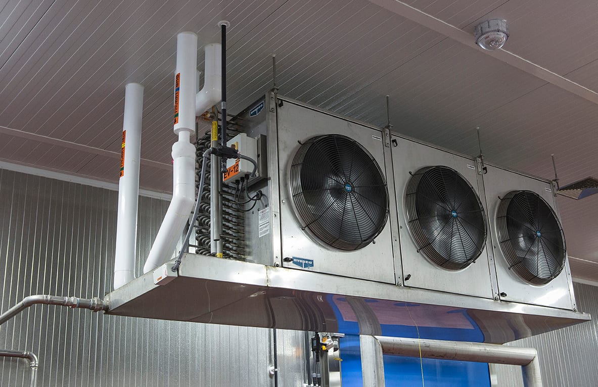 Industrial refrigeration unit with three fans and pipes, suspended from a ceiling in a cold storage facility.