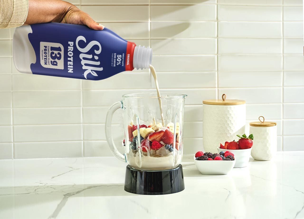 A hand pours Silk Protein milk into a blender with fresh berries and bananas to make a smoothie.