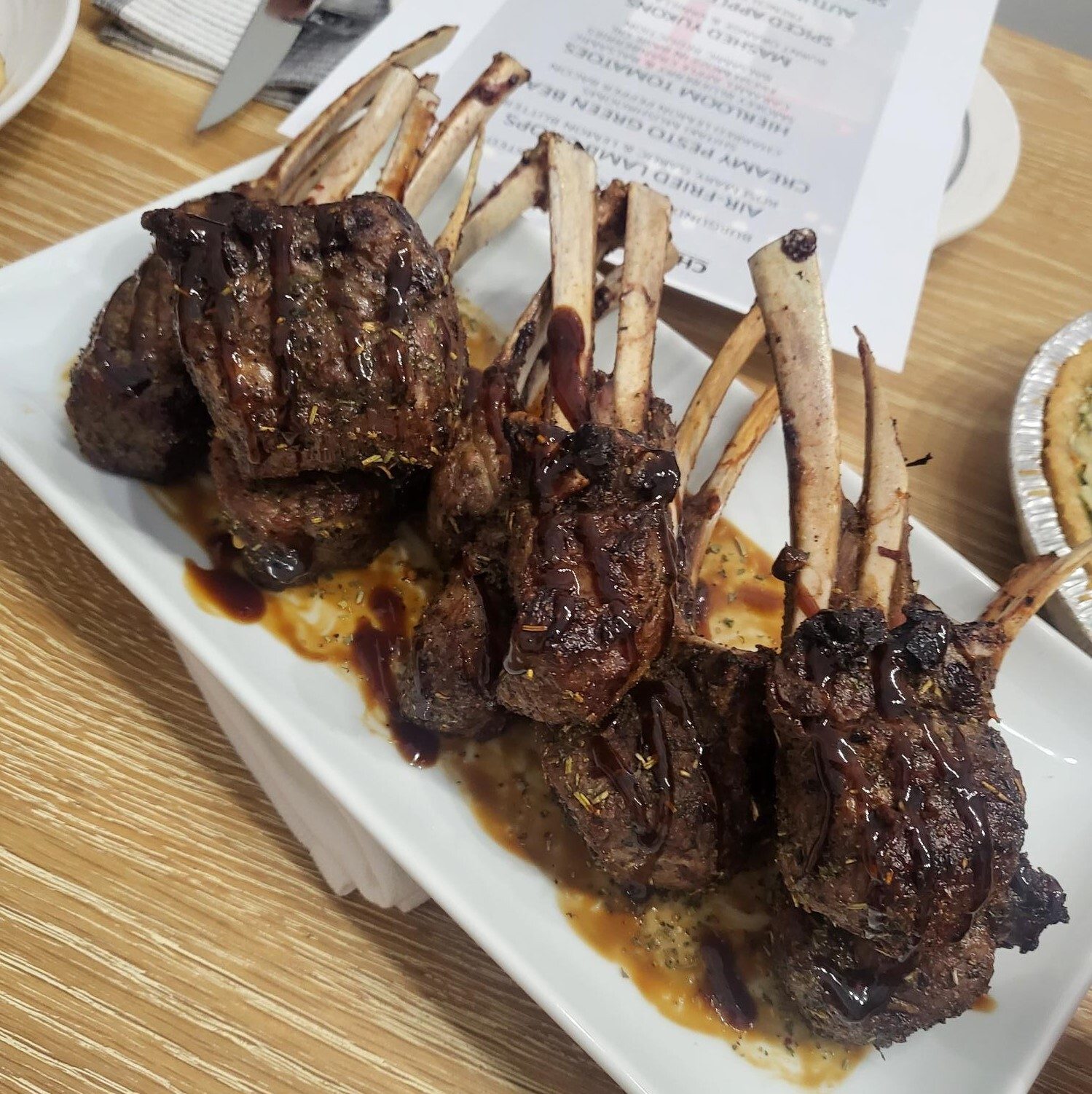 Seasoned lamb chops with bones and dark sauce on a white plate, menu partially visible.