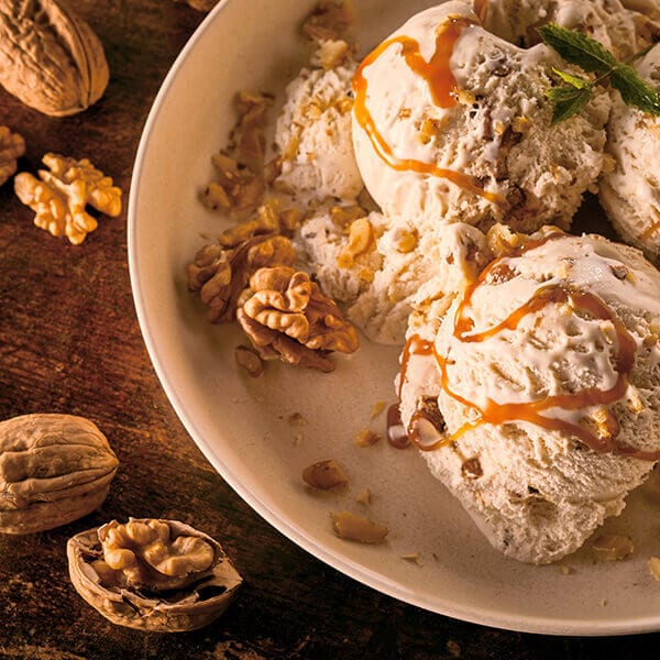 Walnut ice cream with caramel on a plate, whole and chopped walnuts on wood.
