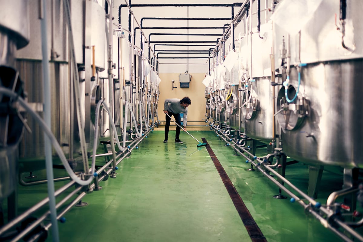 Man cleaning wet green floor in a facility with large stainless steel tanks.