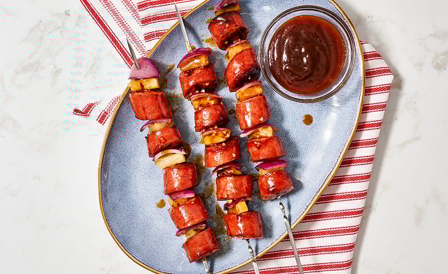Sausage, pineapple, onion skewers with BBQ sauce on a blue plate.