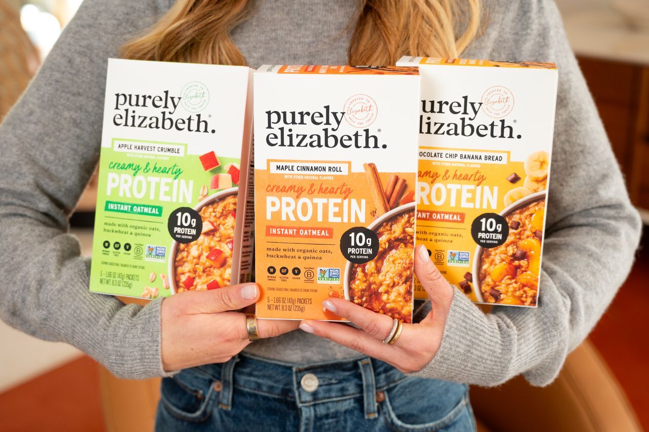 A person holds three Purely Elizabeth instant protein oatmeal boxes in various flavors.