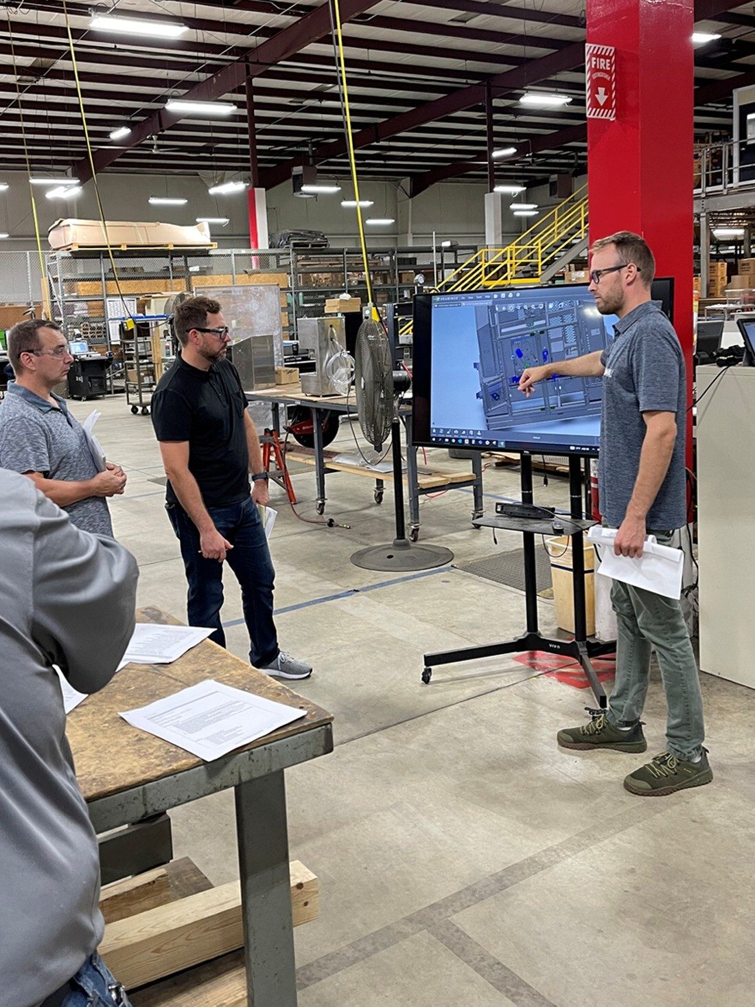 Three men examine a CAD drawing on a large screen in a warehouse, one points at the display.