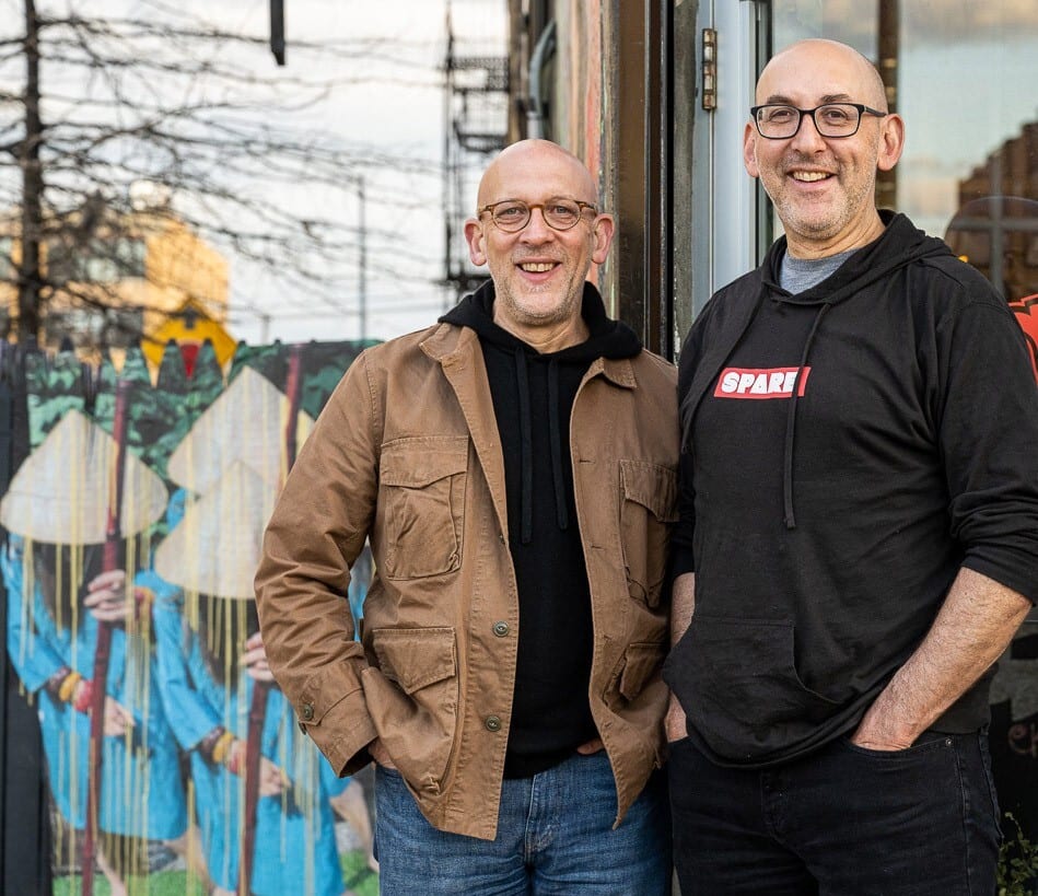 Two smiling bald men in glasses stand outdoors, one in a brown jacket, one in a 'SPARE!' hoodie.