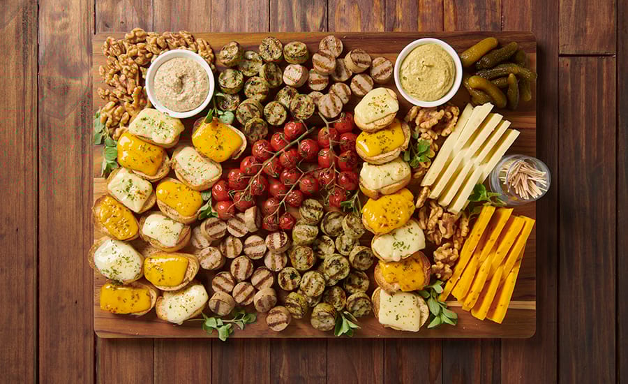 Overhead view of a party platter with grilled meats, cheese, tomatoes, nuts, pickles, and dips.
