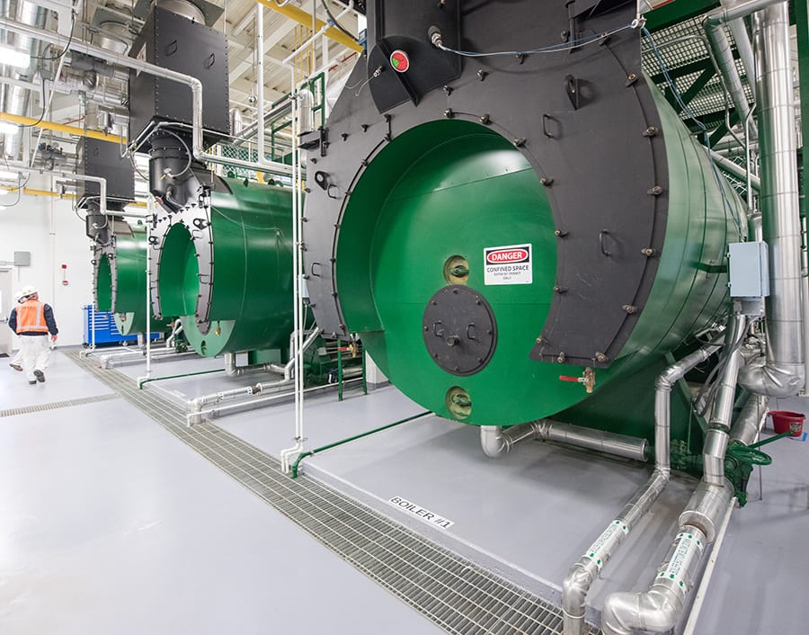 Industrial room with three large green boilers and a worker walking by.