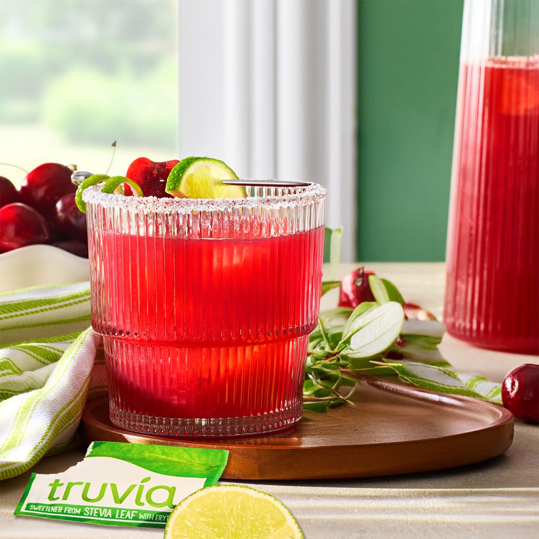 Red drink with lime and sugar rim in a ribbed glass, Truvia packet, pitcher, cherries.