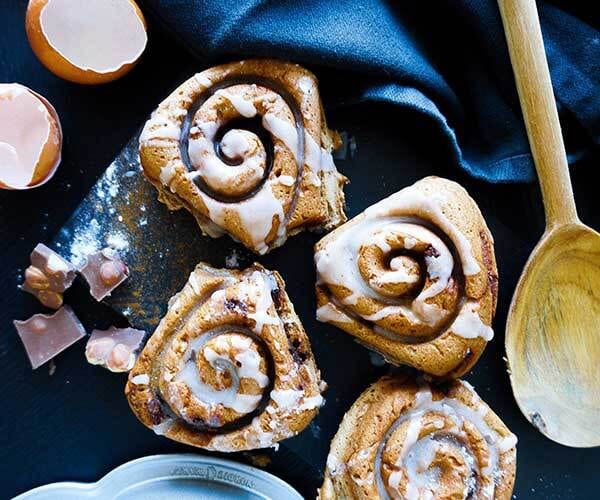 Four iced cinnamon rolls on a dark surface with eggshells, chocolate pieces, and a wooden spoon.