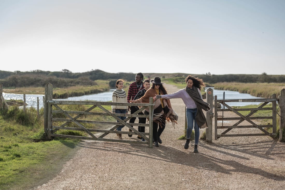 Five diverse people walk through a wooden gate on a sunny outdoor path by water.