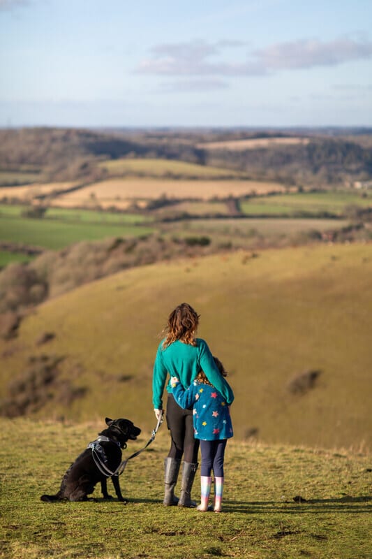 Rear view of a woman, child, and dog on a hill overlooking a vast, green valley.