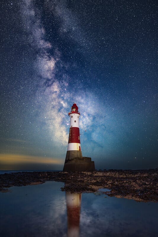 Red and white lighthouse under the Milky Way, reflected in water.