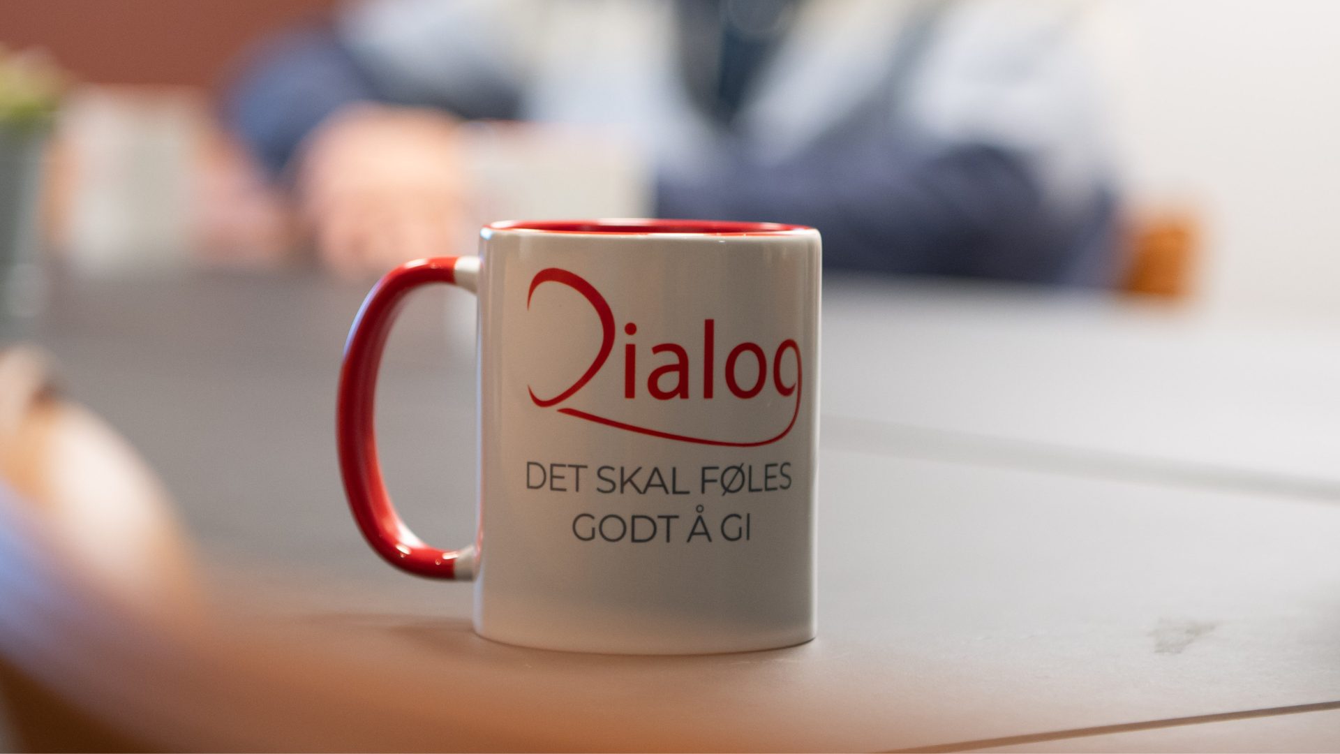 White mug with red handle, "Dialog" logo, and Norwegian text "DET SKAL FØLES GODT Å GI".