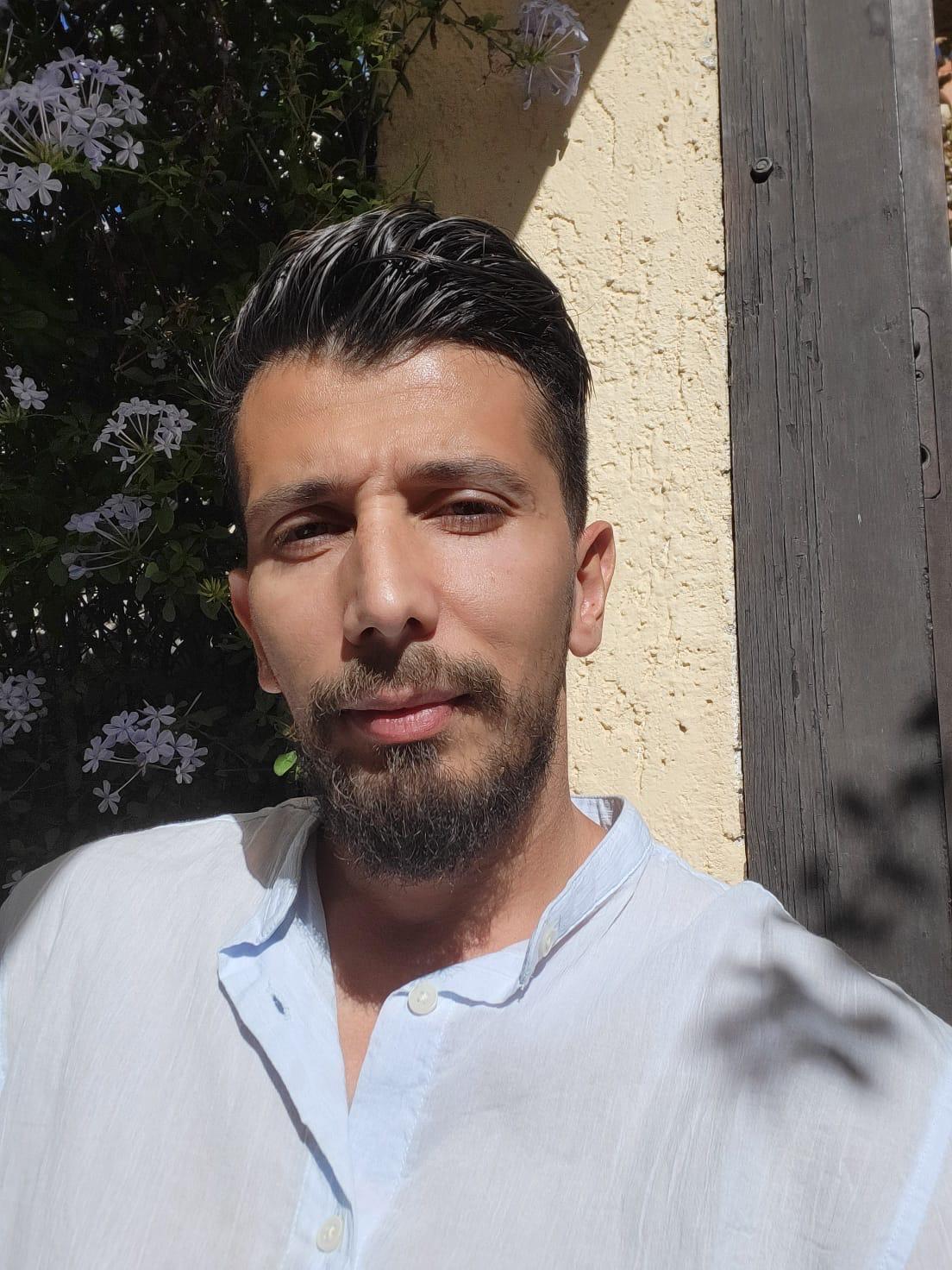 Man with dark hair and beard in a light blue shirt, outdoors with flowers and yellow wall.