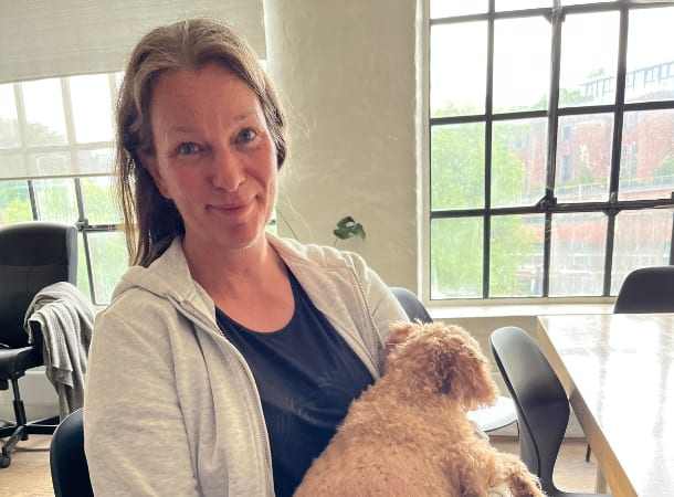 Smiling woman holding a fluffy light brown dog, indoors by a window.