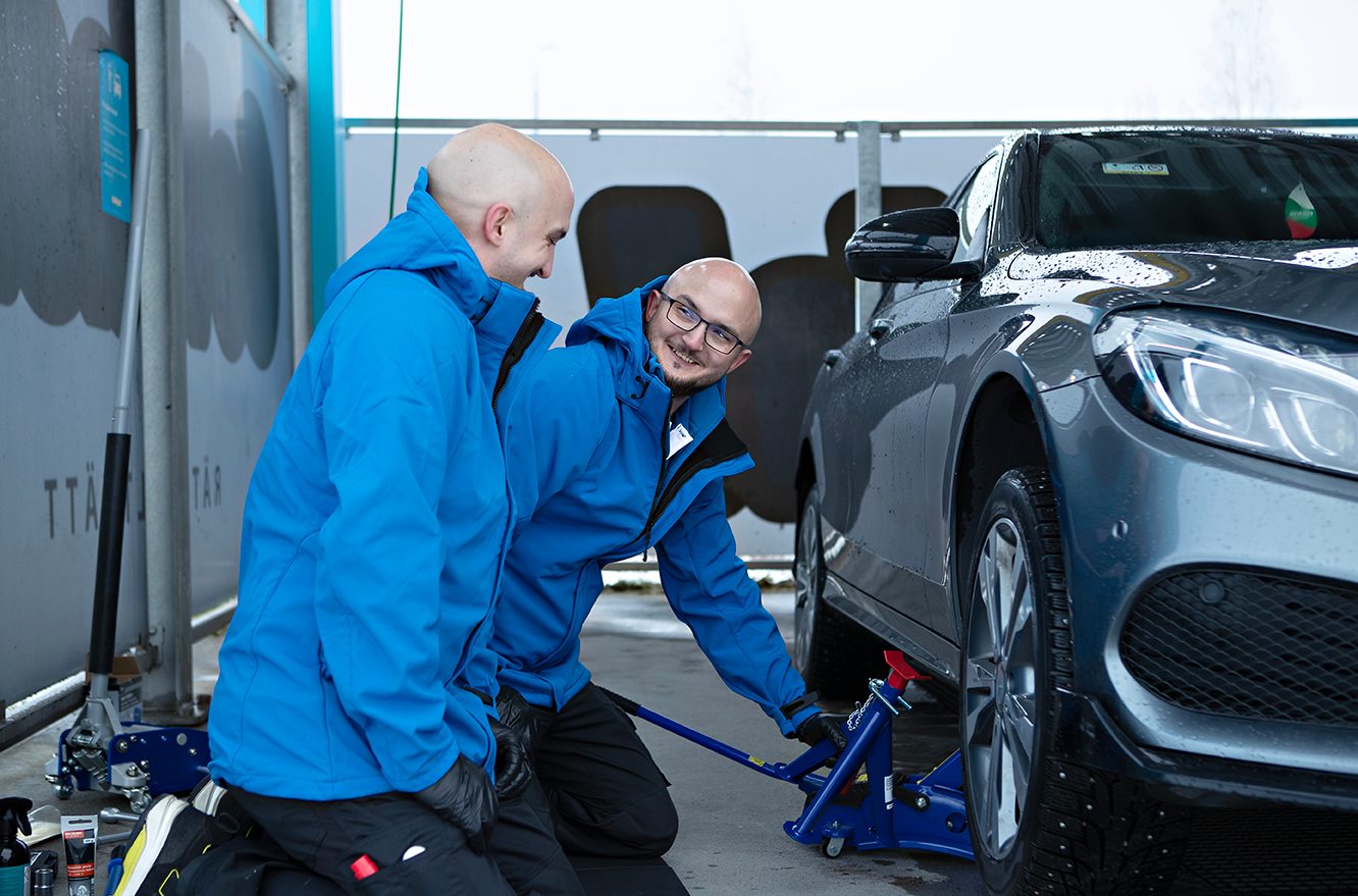 Two bald men in blue jackets using a car jack to lift a dark car, smiling.