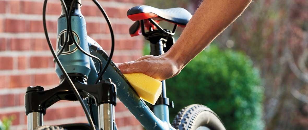 A person's hand using a yellow sponge to clean a teal bicycle frame outdoors.