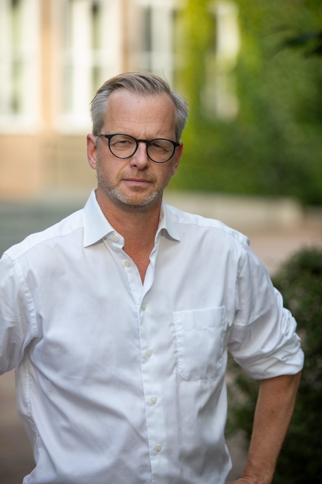 Man in glasses and white shirt looks seriously at camera outdoors with a blurred background.