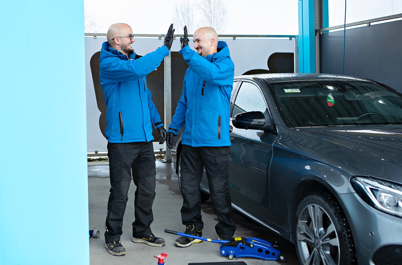 Two auto service technicians in blue jackets high-fiving next to a grey car and a blue car jack.