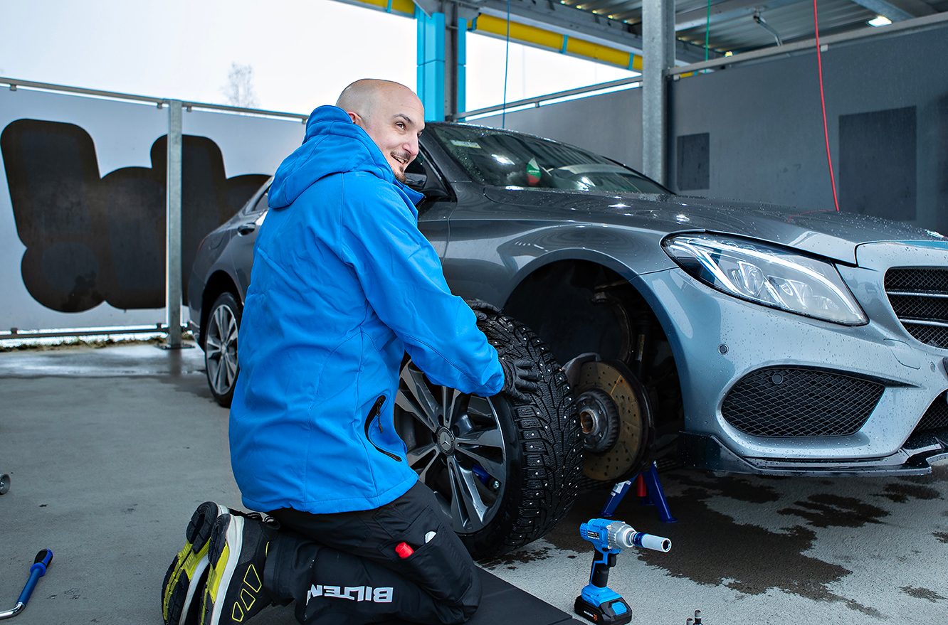 Man kneeling, installing a studded winter tire on a grey car.