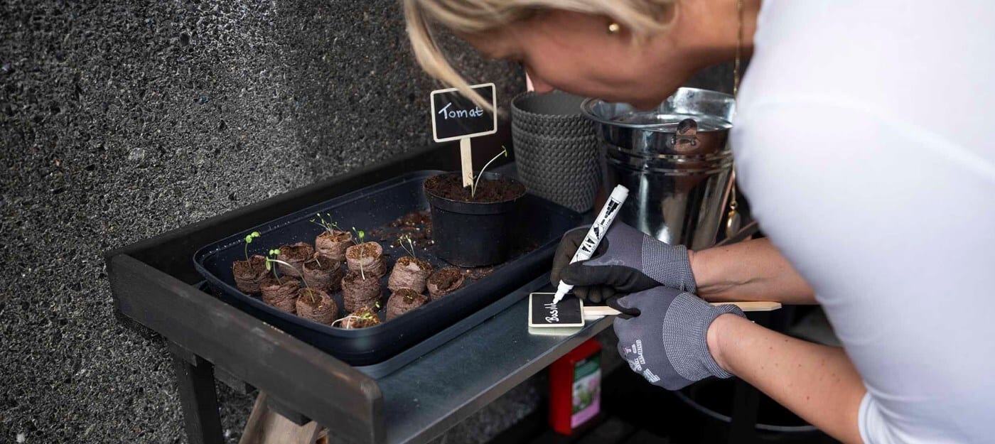 A woman in gardening gloves writes "Basil" on a plant marker, near a tray of seedlings and a potted plant.