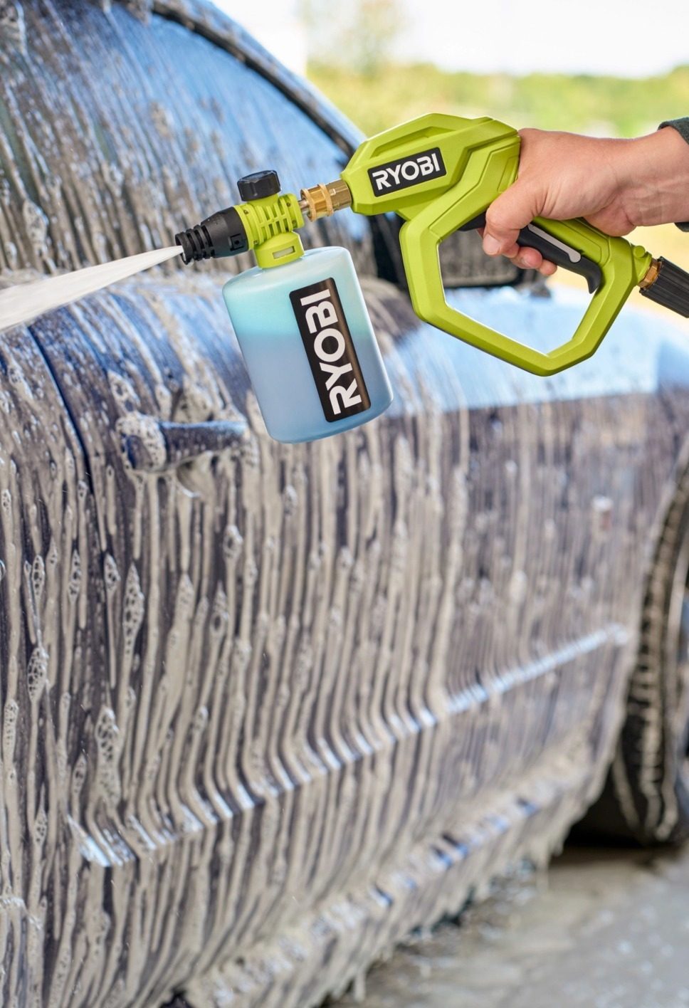 A hand using a green Ryobi foam cannon to spray soap on a car, covering it in white suds.