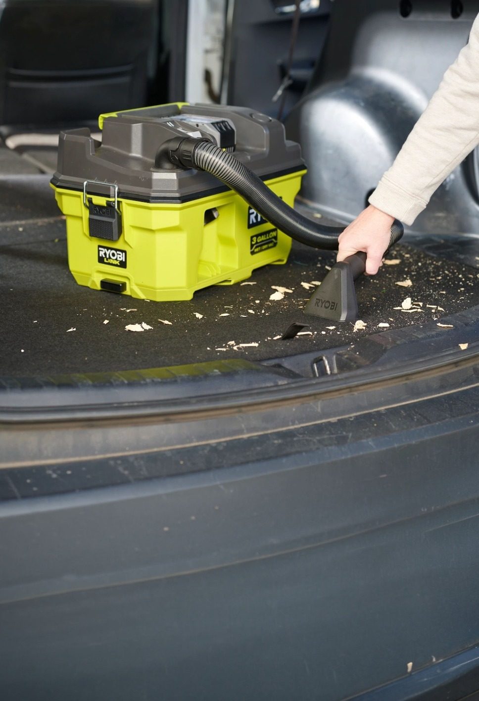 A person uses a green Ryobi portable vacuum to clean sawdust and debris from a car's trunk.
