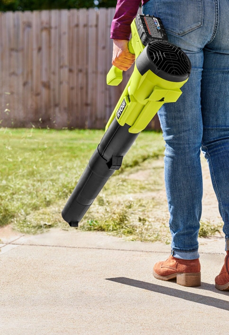 Person in blue jeans holds a green and black Ryobi leaf blower, cleaning a paved area in a garden.