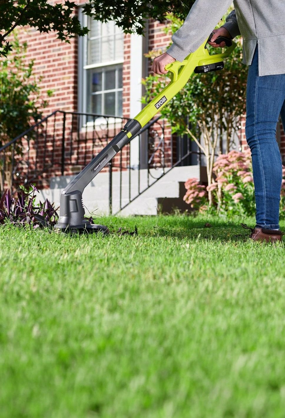 A person uses a green and black Ryobi grass trimmer to edge the lawn in front of a brick house.