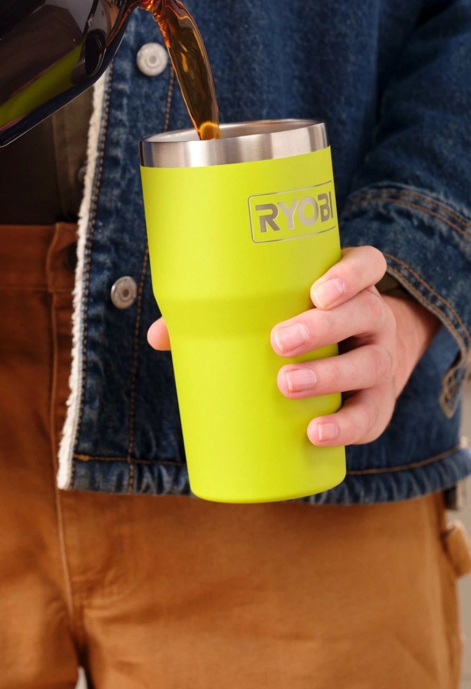 Coffee is being poured into a lime green Ryobi tumbler held by a hand. A person in a denim jacket and brown pants is visible.