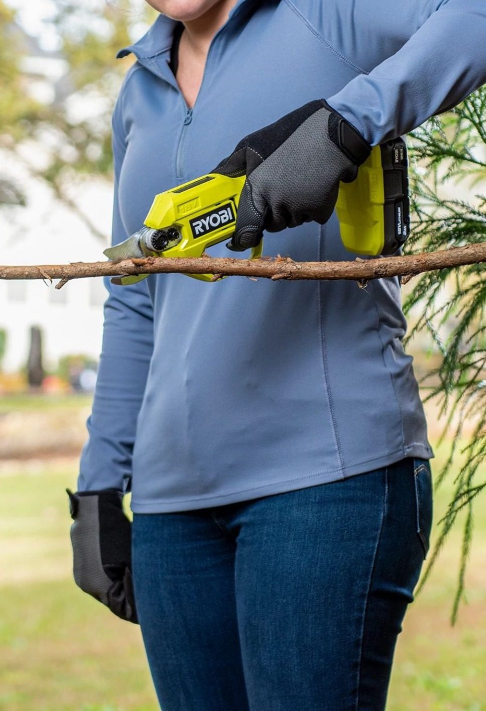 A person in blue uses yellow Ryobi electric pruning shears to cut a tree branch, wearing work gloves.