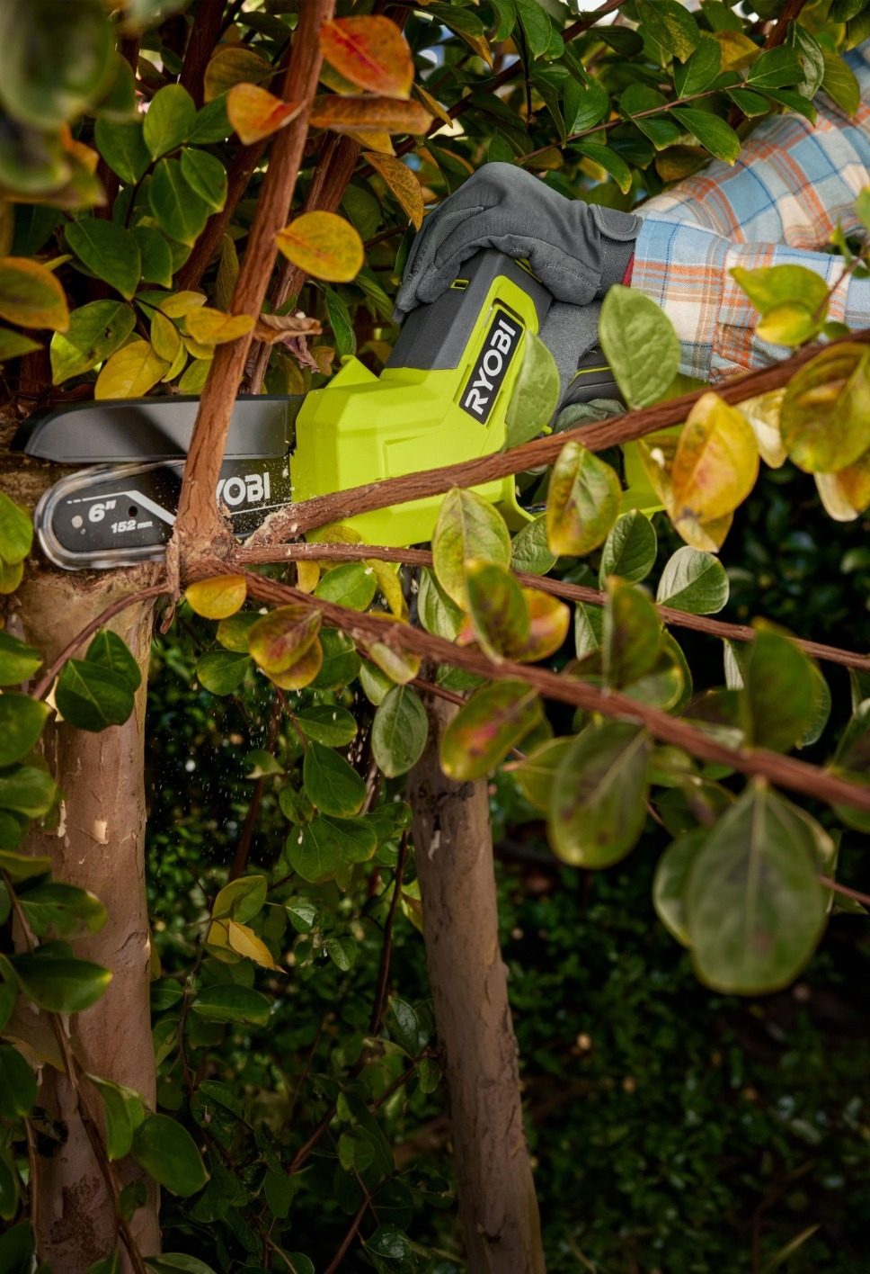 A person uses a lime green Ryobi mini chainsaw to cut a tree branch, with sawdust flying.