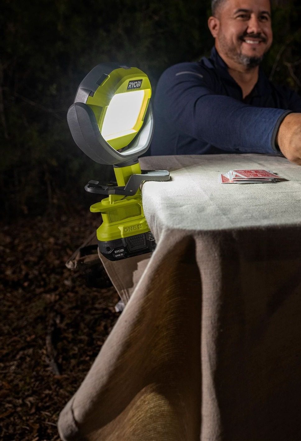 A man plays cards outdoors at night, illuminated by a clamp-on Ryobi work light.