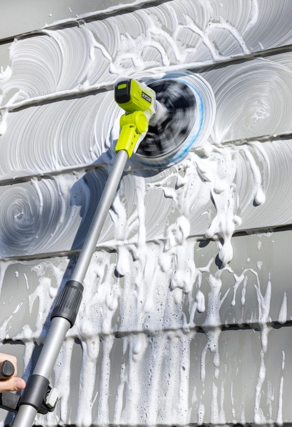 A lime green Ryobi power brush actively cleans grey siding, creating swirling patterns of white soap suds.